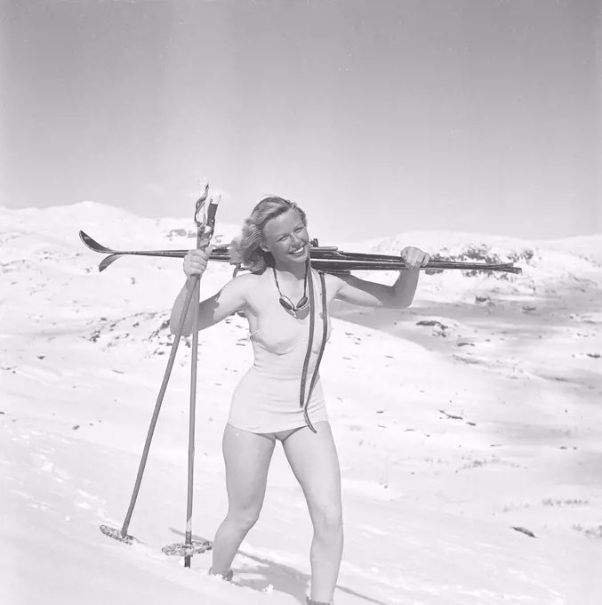 Winter in the 1940s. A young blonde swedish woman is carrying her skis on her shoulder, walking in the deep snow on a mountain top. She is wearing a swimsuit and enjoys the sunny day. Sweden 1940s. Photo Kristoffersson ref H139-6