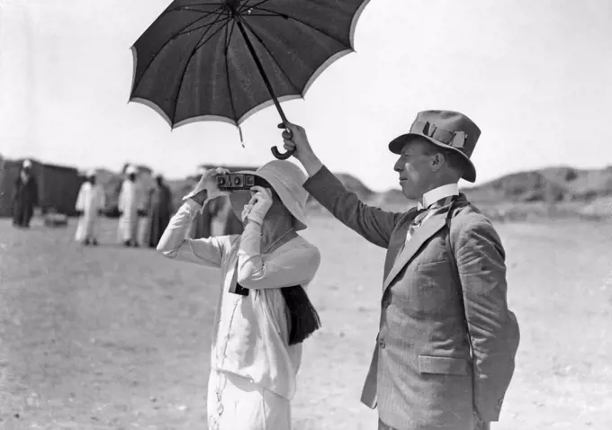 Taking pictures in the 1930s. A woman is photographing something with a Stereo camera. A man is standing by her side holding an umbrella to block the sun. 1930s