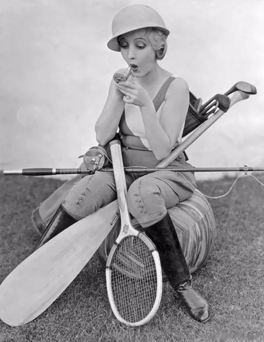 Bessie Love, 1898-1986. American actress pictured here with a lipstick and a small pocket mirror. She is surrounded by different kinds of sport equipment, tennis racket, fishing rod, golf clubs and a kayak paddle. 1930s