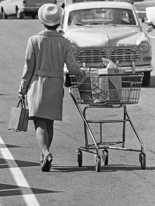 Shopping in the 1970s. A lady is leaving the supermarket and heading for her car pushing the shopping cart on the parking lot. Sweden 1970