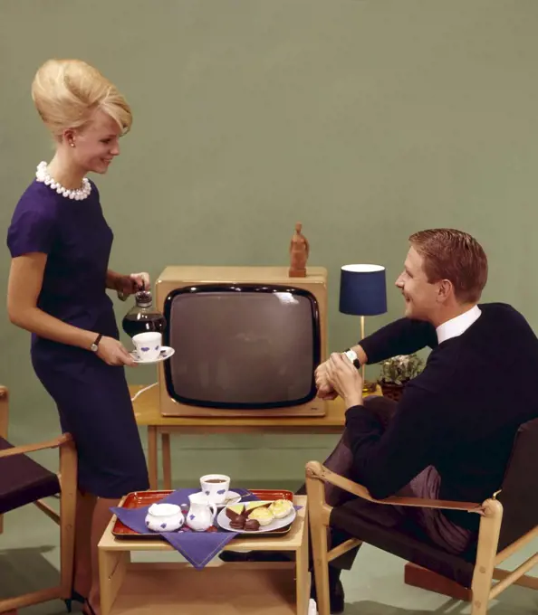 Coffee in the 1960s. A woman is pouring coffee for her husband in front of the tv set. She has her hair in the typical Beehive hairstyle, in which her long hair is piled up on the top of the head and giving some resemblance to the shape of a traditional beehive. The chairs are of the Safari design type. Sweden 1960s