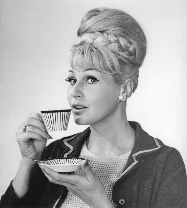 Coffee in the 1960s. A woman is drinking coffee from a coffeecup with a 1960s striped pattern. She has her hair in the typical Beehive hairstyle, in which long hair is piled up on the top of the head and giving some resemblance to the shape of a traditional beehive. Sweden 1962