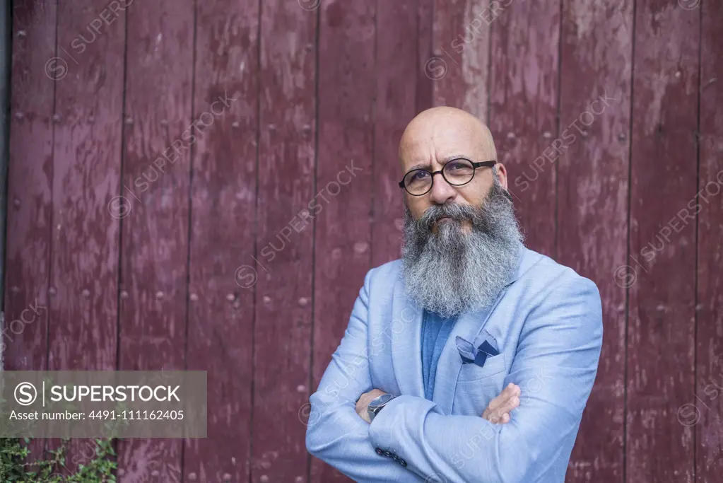 Front view of a mature trendy bearded hipster standing against a vintage red door outdoors while looking to camera