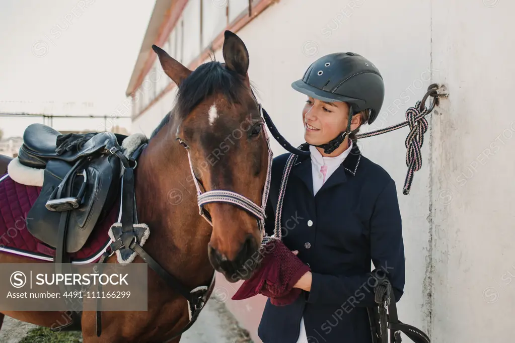 Side view of young teen woman in jockey helmet and jacket caressing horse standing together outdoors