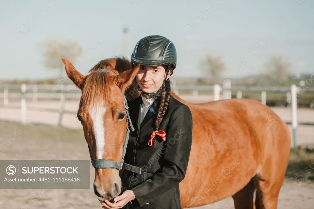 Side view of young teen woman in jockey helmet and jacket caressing horse standing together outdoors and looking at camera