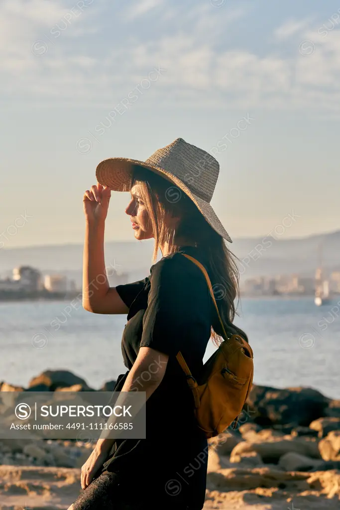 Side view of female tourist wearing a straw hat and backpack standing near beach