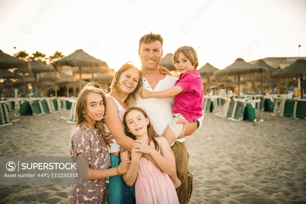 Adult loving man and woman with son and daughters standing together on beach in back lit smiling at camera