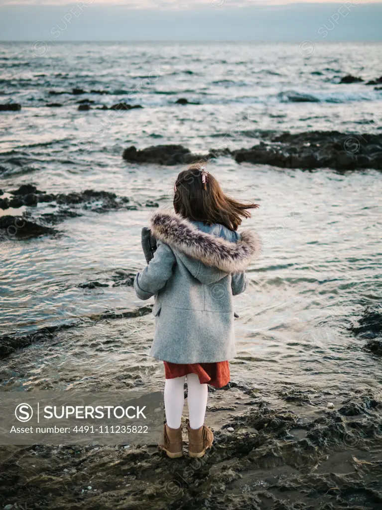 anonymous little girl from behind standing by the beach on rocks looking at the sea on a winter day