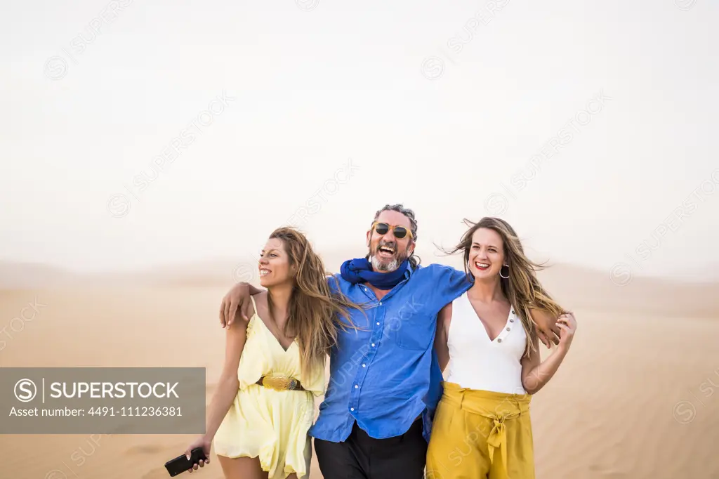 Senior bearded man laughing and hugging cheerful women while walking in sandy desert during trip in Morocco