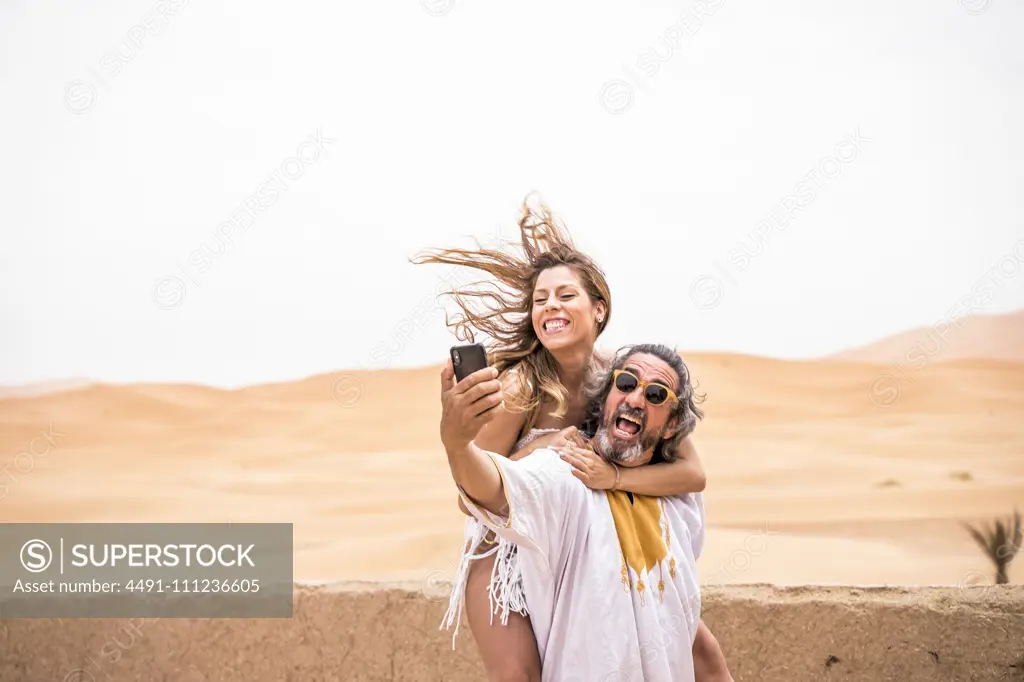 Middle-aged man with woman piggyback taking selfie expressively on terrace against sandy desert, Morocco