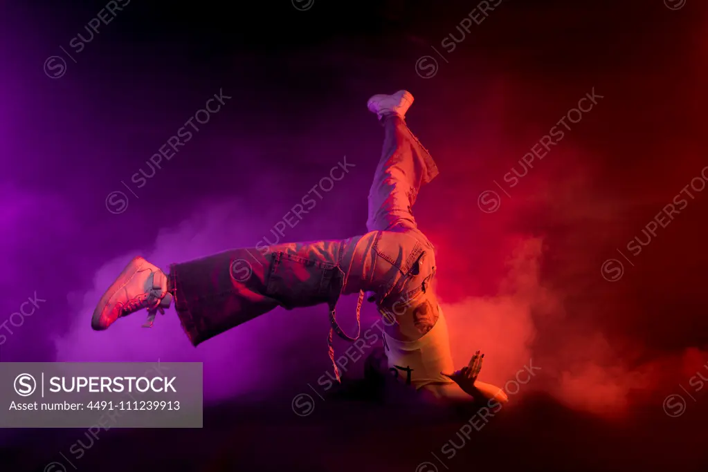 Modern female performing break dance on floor in purple and red smoke on black background