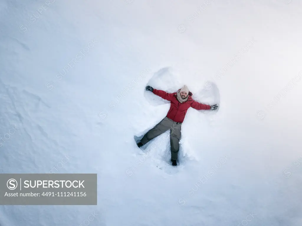 From above smiling middle-aged male in outwear lying on white snow and making snow angel silhouette