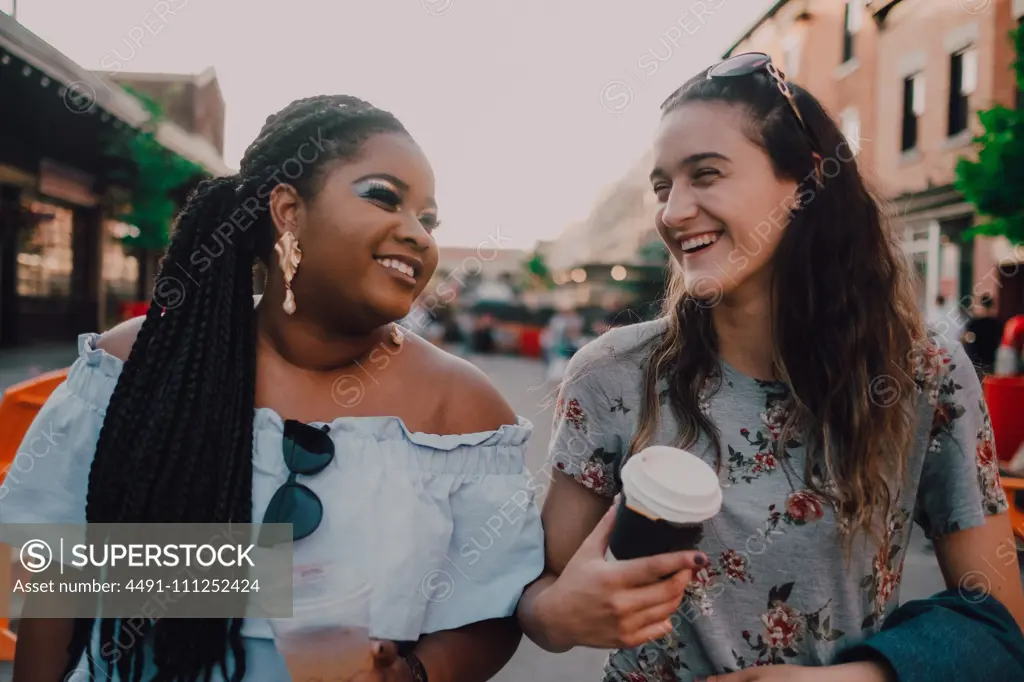 Cheerful trendy multiracial young casual women talking and drinking coffee while walking on street on sunset