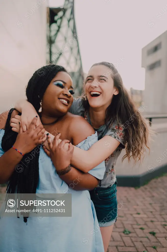 Playful multiracial young casual women laughing and hugging while standing on street on sunset