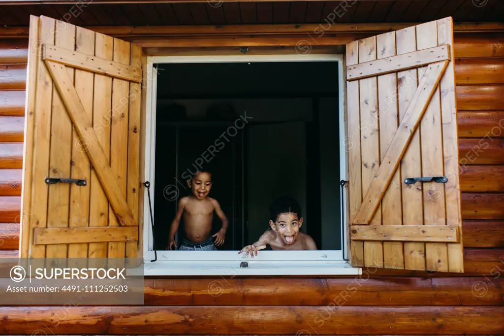 Two shirtless African American brothers looking out open window of wooden house while having fun at home together