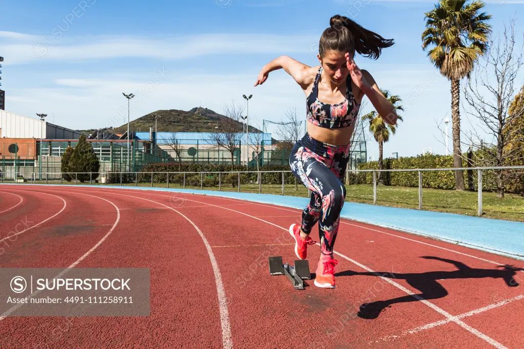 Strong female athlete in sportswear running fast against blue sky on sunny day on stadium