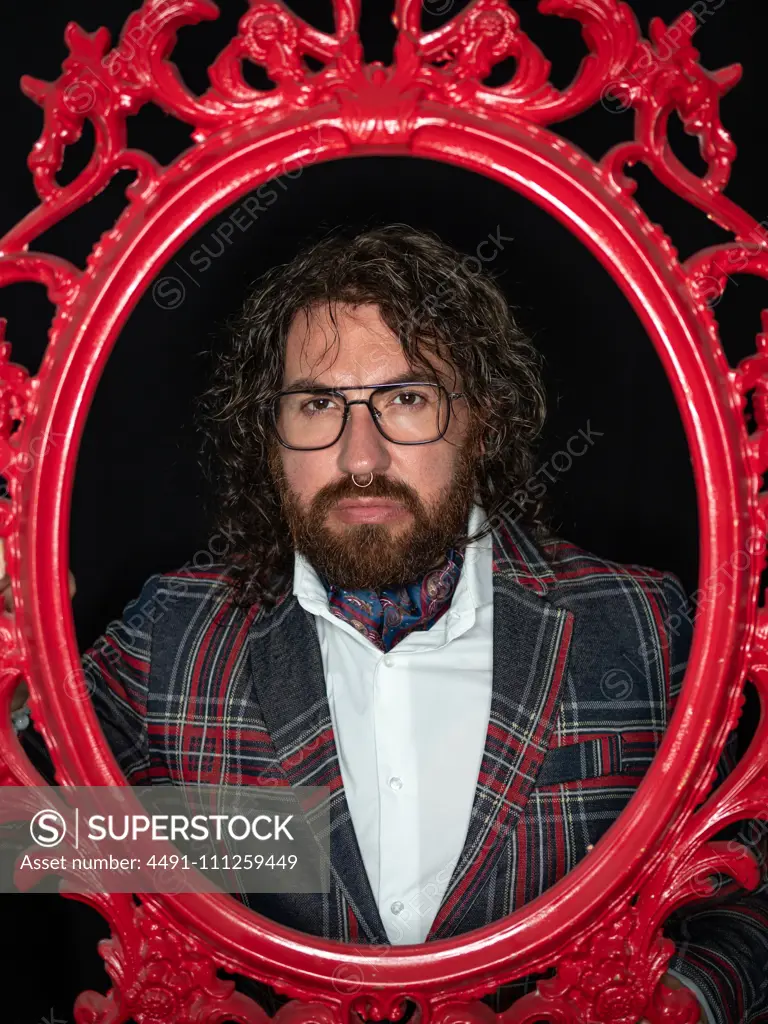 Serious impressive well dressed man with curly hair and beard posing in red patterned frame on black background