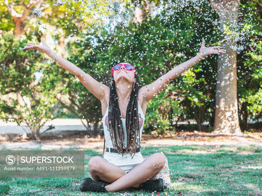 Joyful young female with dreadlocks sitting on ground legs crossed throwing colorful confetti and looking up