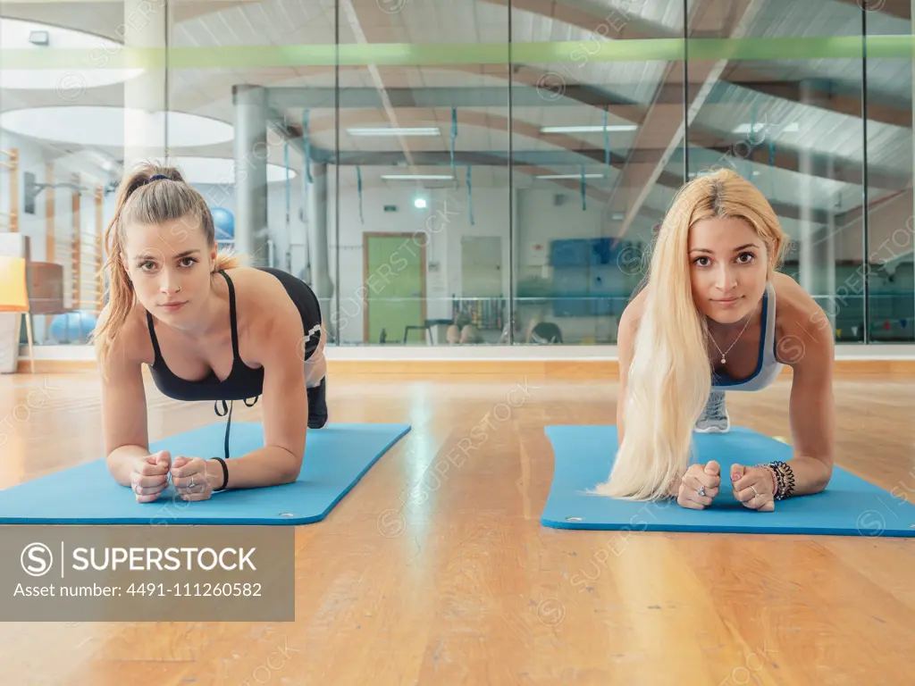 Two muscular slim women in sportswear looking at camera while doing plank on mat in gym