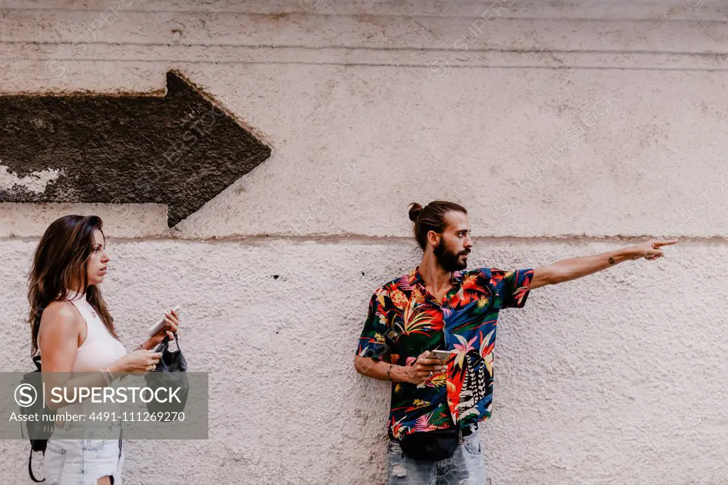 Handsome young bearded man showing path to thoughtful brunette with backpack and smartphone against white wall with arrow