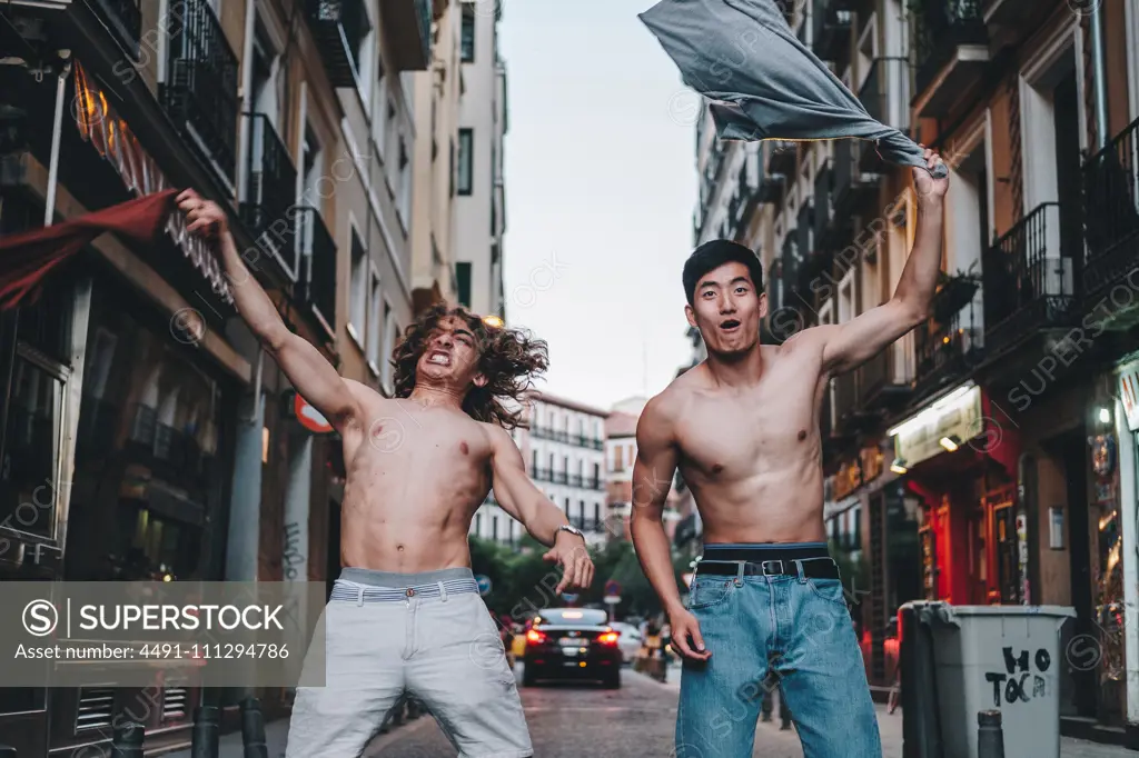 Screaming multiethnic men taking off shirts and waving above head while standing on urban summer street