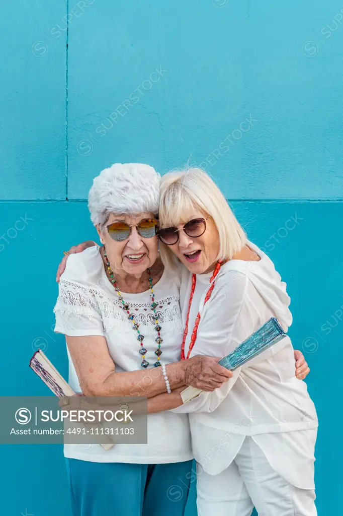 Positive trendy aged grey-haired females waving with big color hand fans talking looking at each other on blue background in summer outside