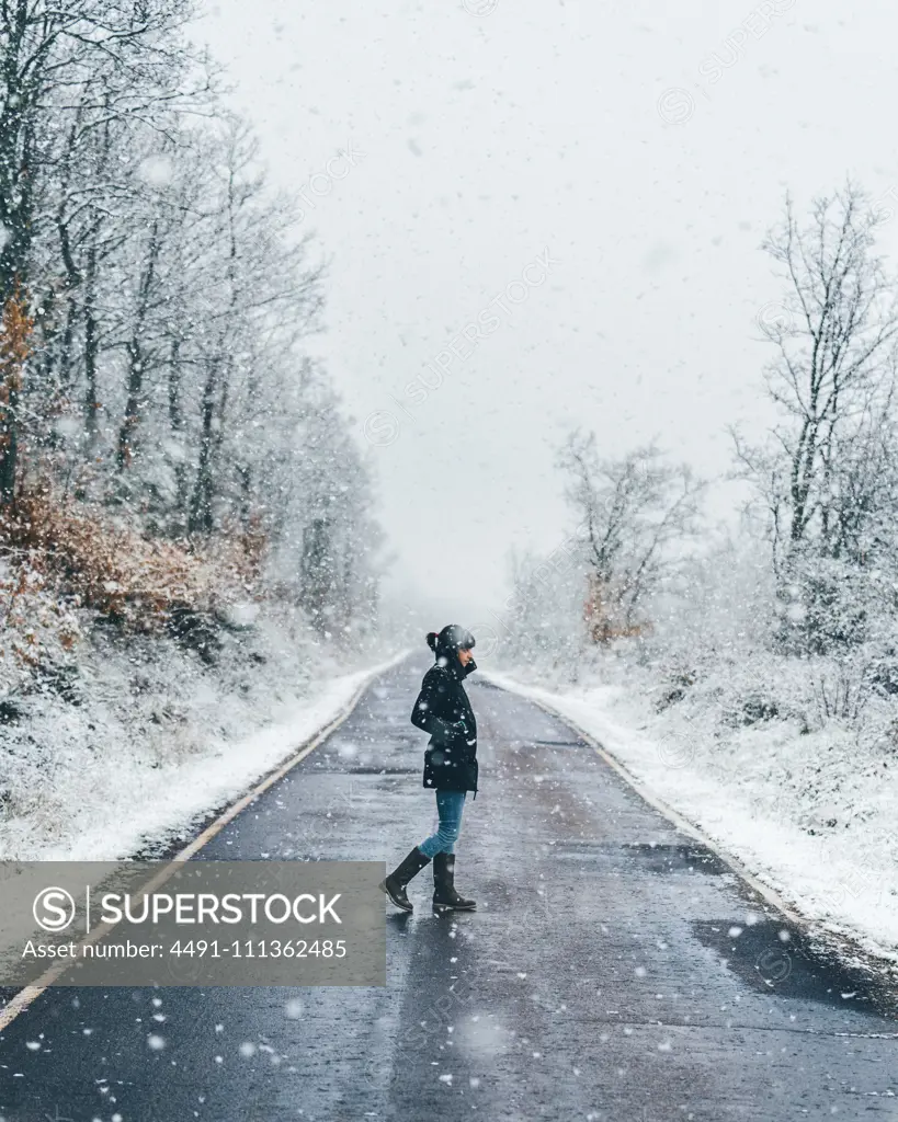 Side view of lonely female in black jacket and jeans crossing empty country road on snow and gloomy winter day