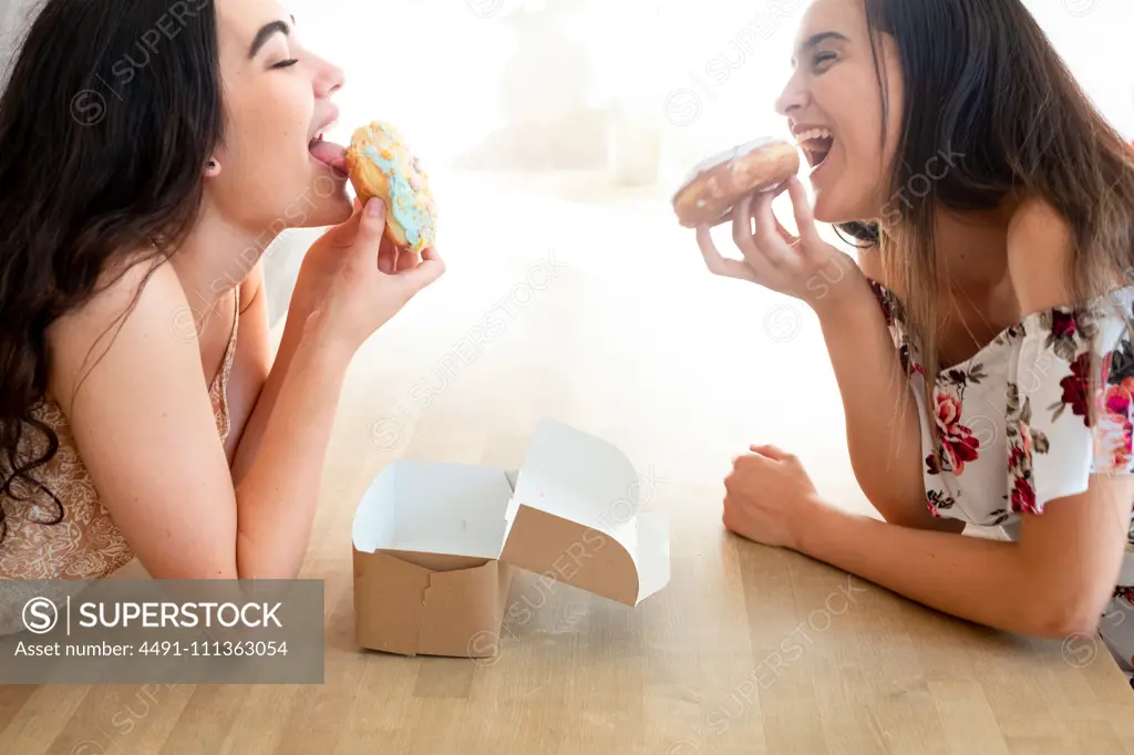 Happy women in sundresses with box of glazed desserts while sitting at table