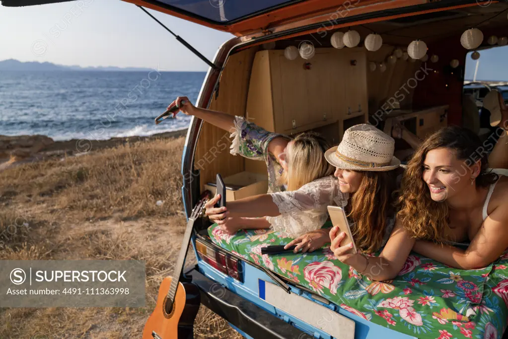 Cheerful pleasant ladies lying on trunk of bright minivan and having fun taking selfie on mobile phone on beach in sunny day