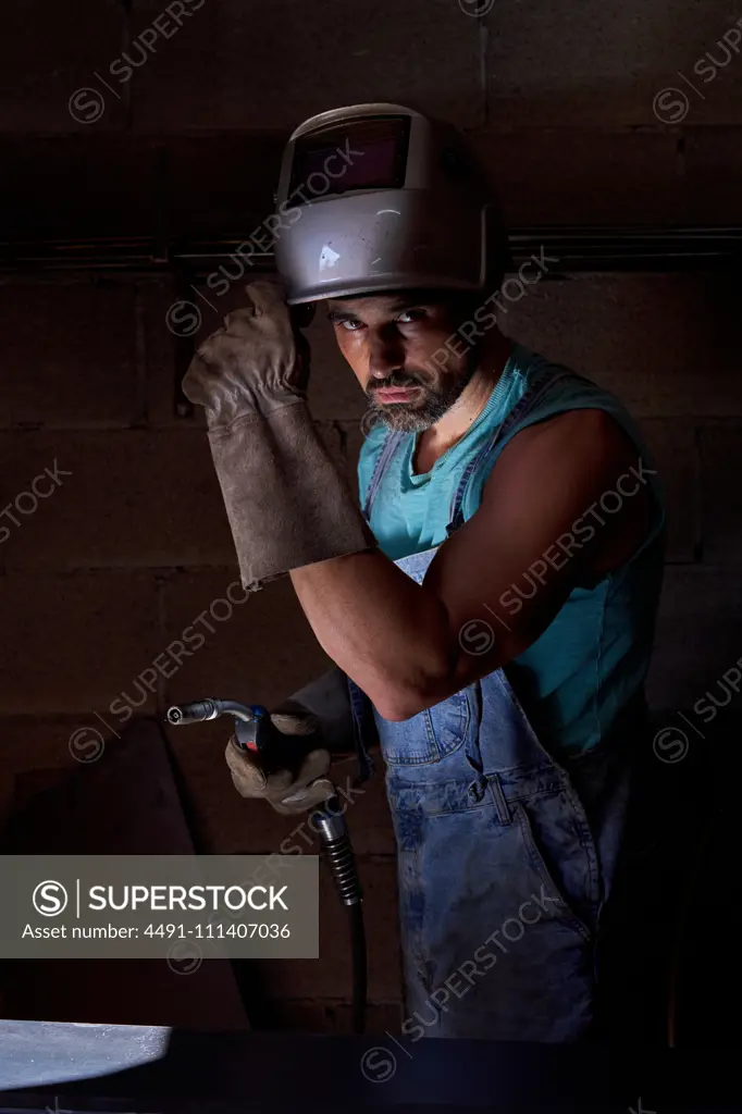 Professional man with welding mask on head wearing jeans overalls and  protective gloves standing on workplace and preparing instrument for  soldering while looking at camera - SuperStock