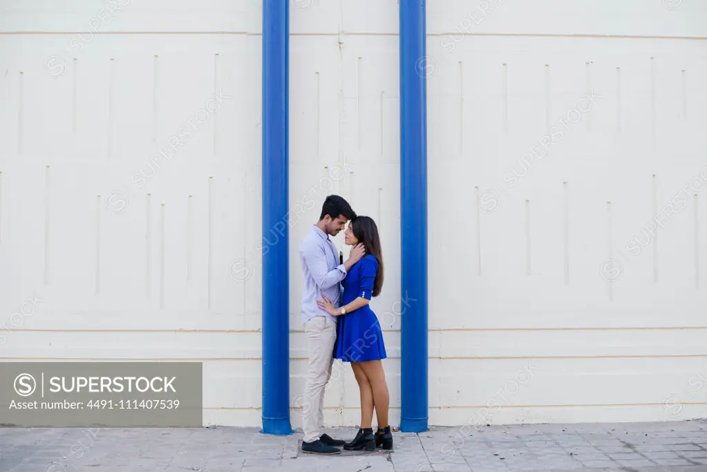 Side view of loving man and woman in elegant clothes kissing on background of white wall with bright blue columns