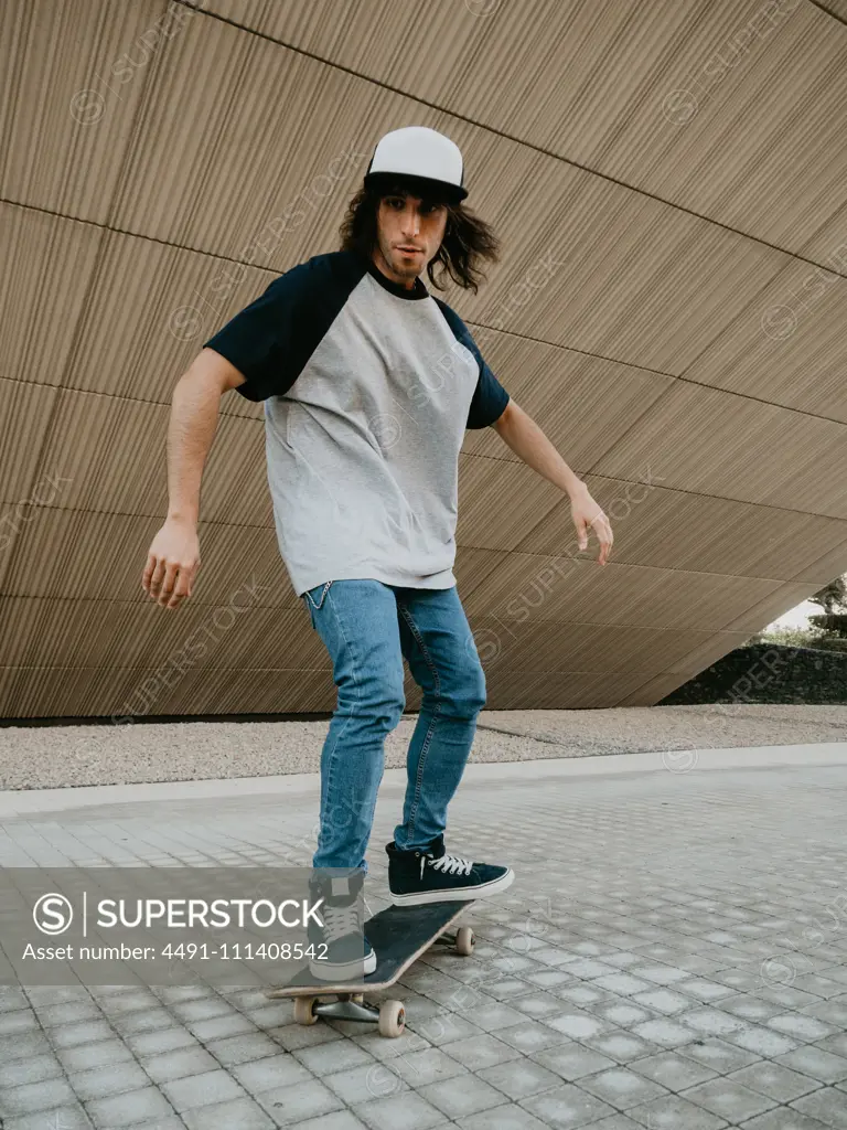 Cool millennial guy in cap balancing while riding skateboard on city street under sloping wall