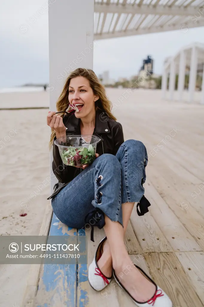 Stylish long haired blonde woman in black jacket enjoying healthy green salad while sitting on wooden terrace on coast smiling looking away