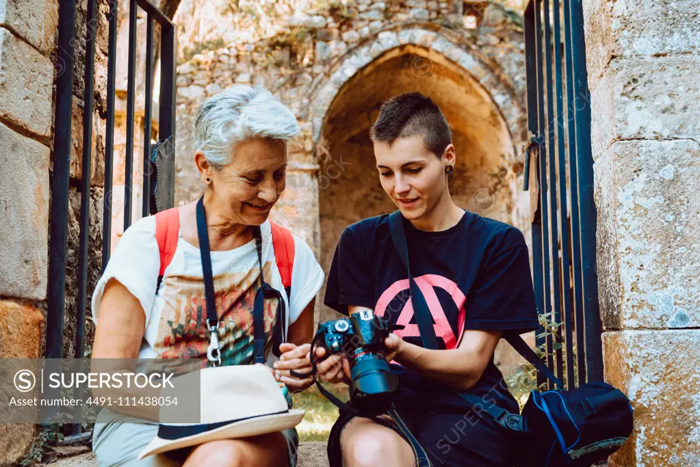 Enthusiastic grandmother and granddaughter sitting at stony entrance of ancient building with metal doors and watching photo pictures on camera