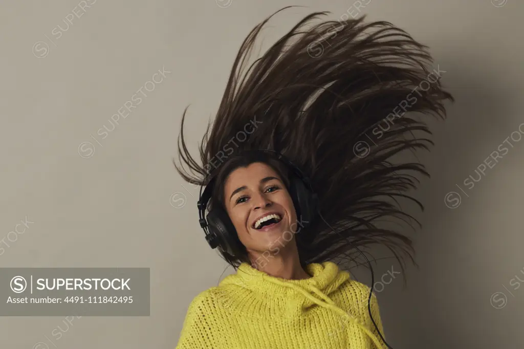 Amazed youthful attractive female brunette in yellow sweater and headphones looking at camera and jumping while listening music on gray background in studio