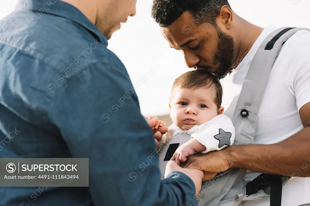 Side view of black casual man holding hand of anonymous male boyfriend and holding little calm baby in grey carrier while strolling on nature at daytime