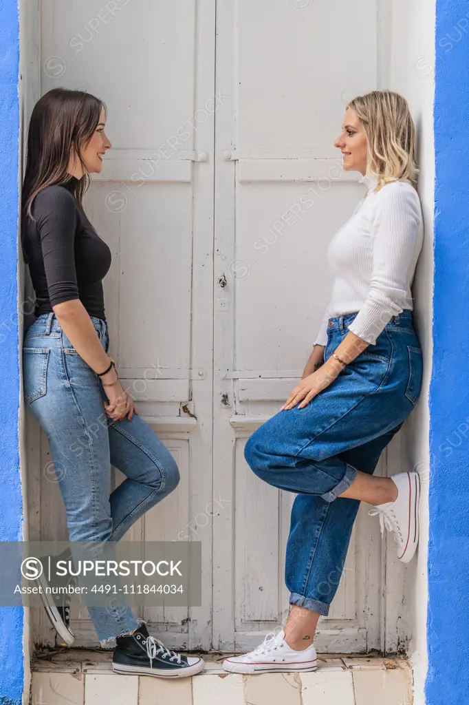 Side view of happy gentle caring girlfriends in stylish outfit standing near door looking at each other