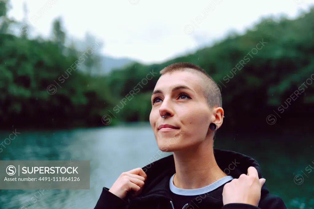 Young extraordinary woman with short hairstyle and piercing in casual clothing looking up to sky with pond among green plants on blurred background