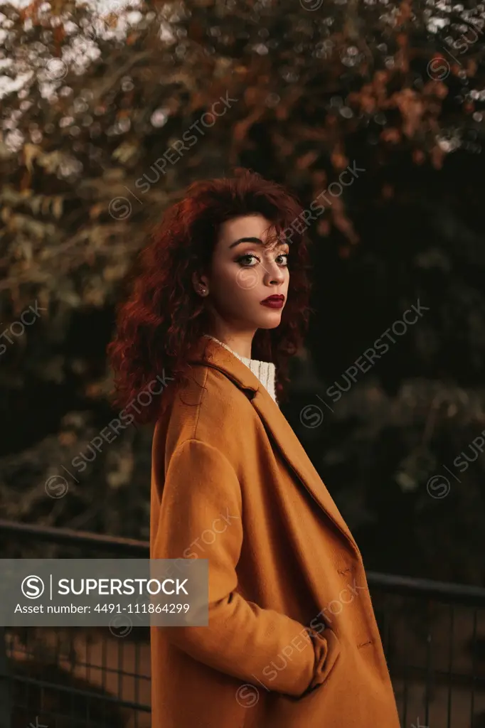 Side view of young sensual female with dark curly hair wearing knitted jumper and overcoat standing in park putting hand on metal railing while looking at camera with thoughtful look