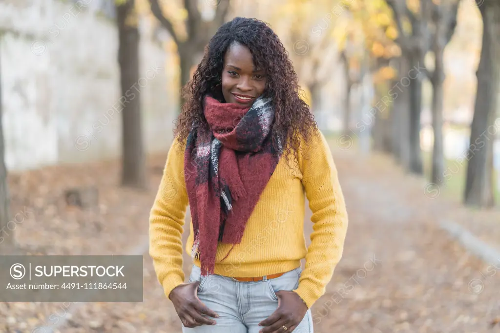 Charming fashion African American woman in a winter scarf with hands in pocket on road with autumn leaves in park