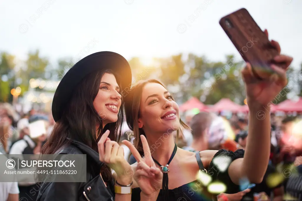 Charming cheerful friends in black hat having fun grimacing and taking selfie on mobile phone in bright day at festival
