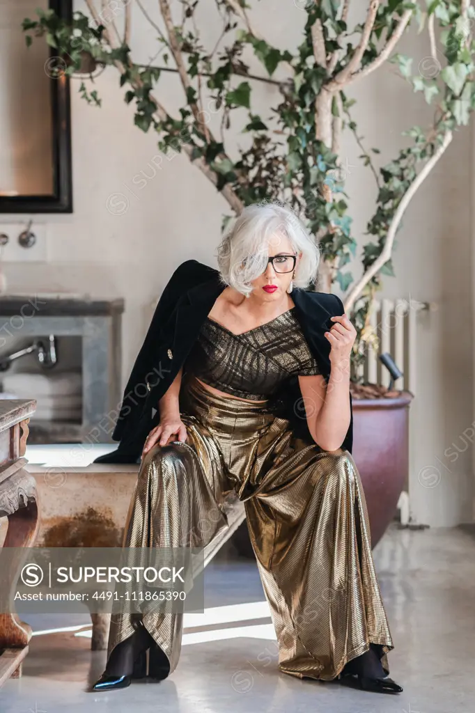 Calm senior confident lady in glasses and elegant clothes looking at camera while sitting on shabby bathtub edge among rich retro interior including old furniture and big green plotted tree in spacious room of country house