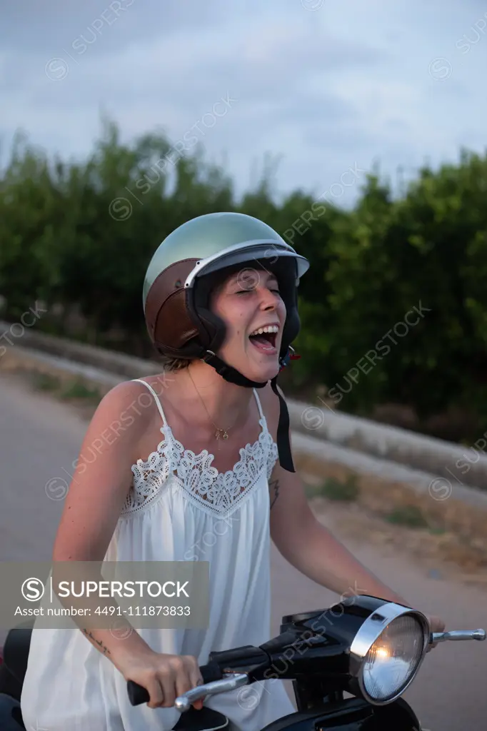 Cheerful woman on vacation in light clothing smiling with closed eyes on motorcycle in motion on the road