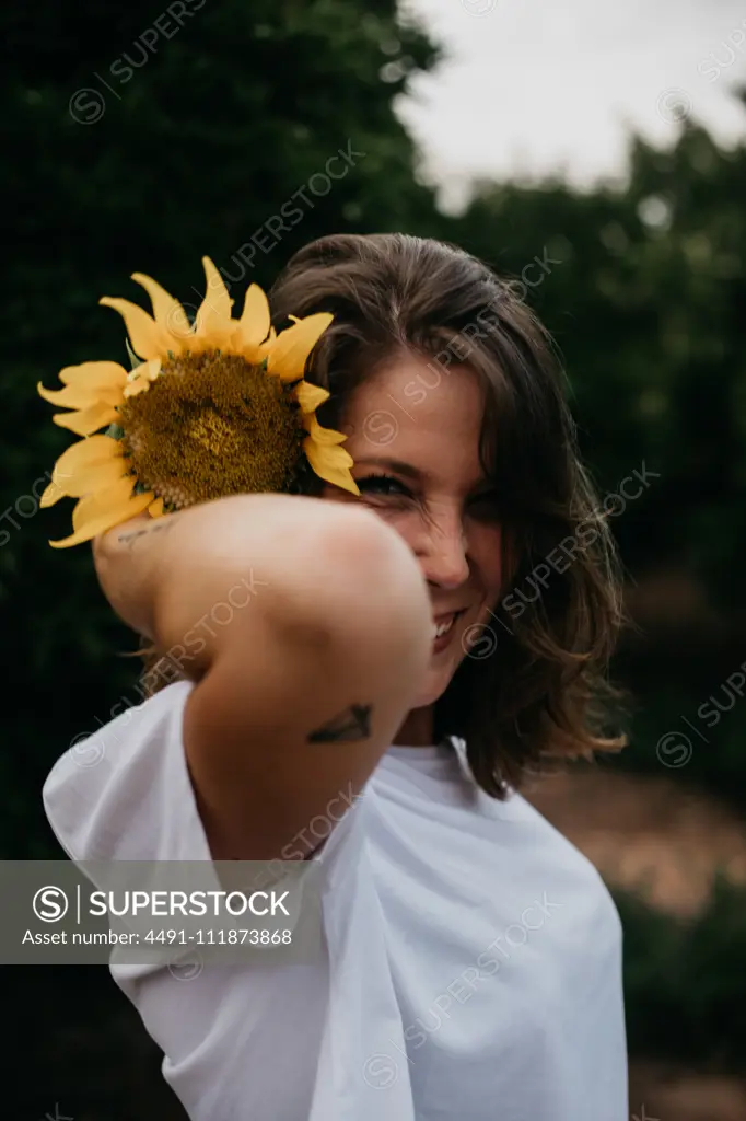 Side view of brown haired woman in light clothing looking at camera holding sunflower behind ear amid green trees during summer vacation