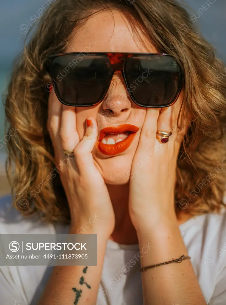 Stylish brown curly haired woman with red lipstick in trendy sunglasses looking at camera while squeezing face in palms;Contemporary resting lady touching face