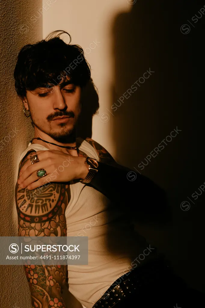 Young stylish guy with mustache and tattoos wearing white tank top and leaning against wall with back while looking away