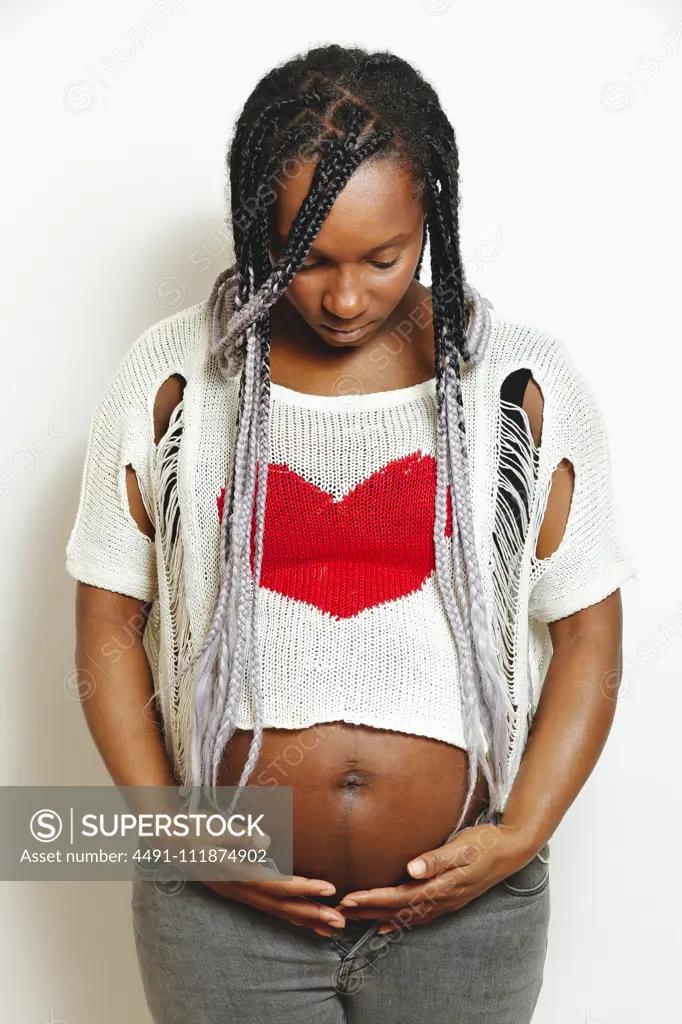 Cheerful young pregnant black female with braids wearing stylish white blouse with red heart touching belly and looking away with smile while standing against white wall