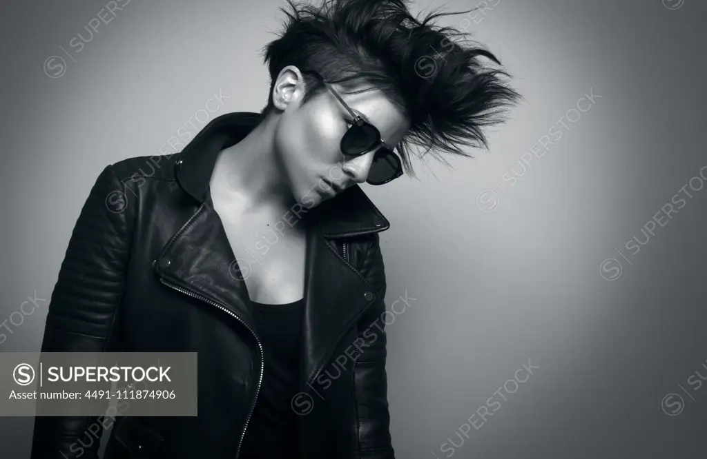 Independent serious youthful woman with short dark hair and sunglasses wearing biker jacket and looking down in studio on gray background;Strong young female rocker in leather jacket in studio