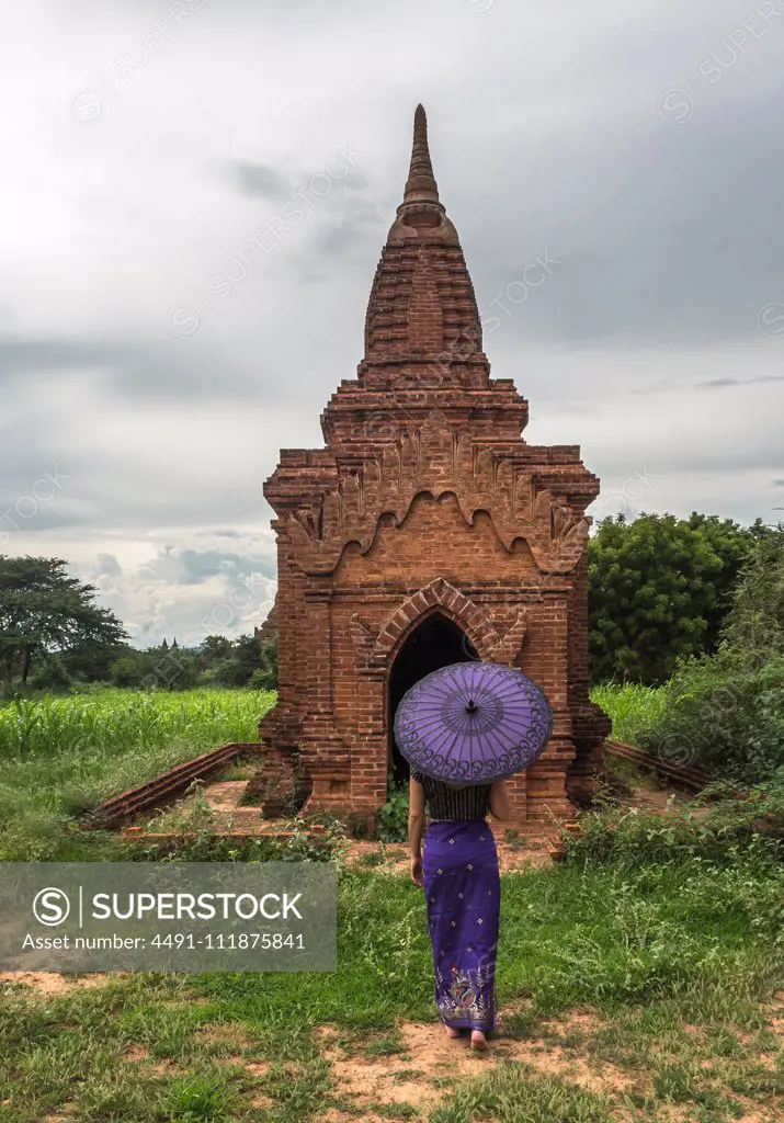 Back view of female in long dress holding traditional Burmese purple umbrella while standing on green meadow in front of old stone temple in ancient city Bagan in cloudy day;Woman with umbrella standing in front of ancient temple