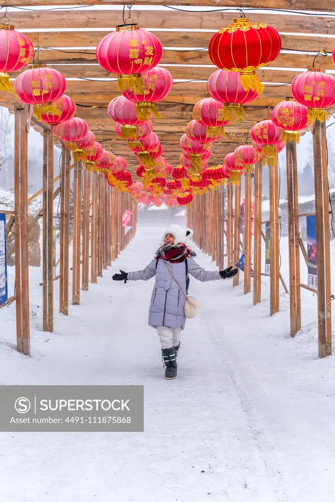 Full body happy young female with outstretched arms admiring red Chinese lanterns while walking in wooden passage on winter day;Cheerful woman walking in archway with Chinese lanterns